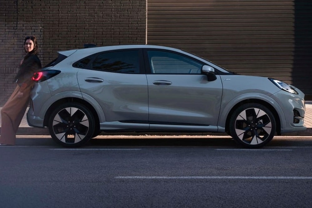 Une Ford Puma stationnée sur la voie publique, avec une jeune femme adossée à la voiture.