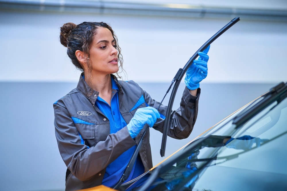 Un technicien Ford, ganté de bleu, remplace un essuie-glace sur le pare-brise d'une voiture dans un garage.