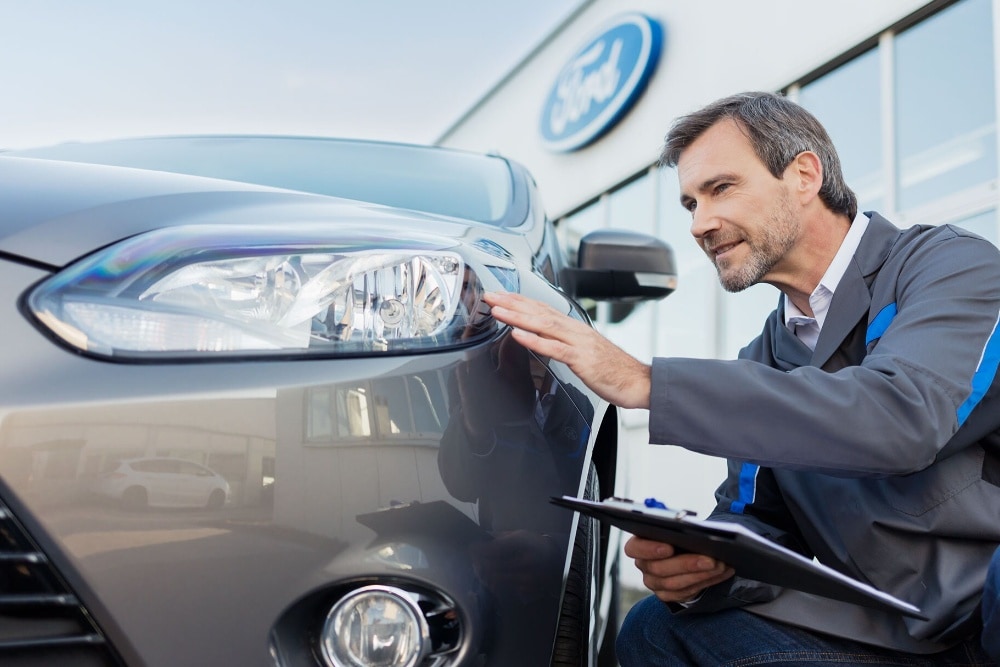 Un technicien Ford inspecte le phare d'une voiture devant une concession Ford, un presse-papiers à la main.