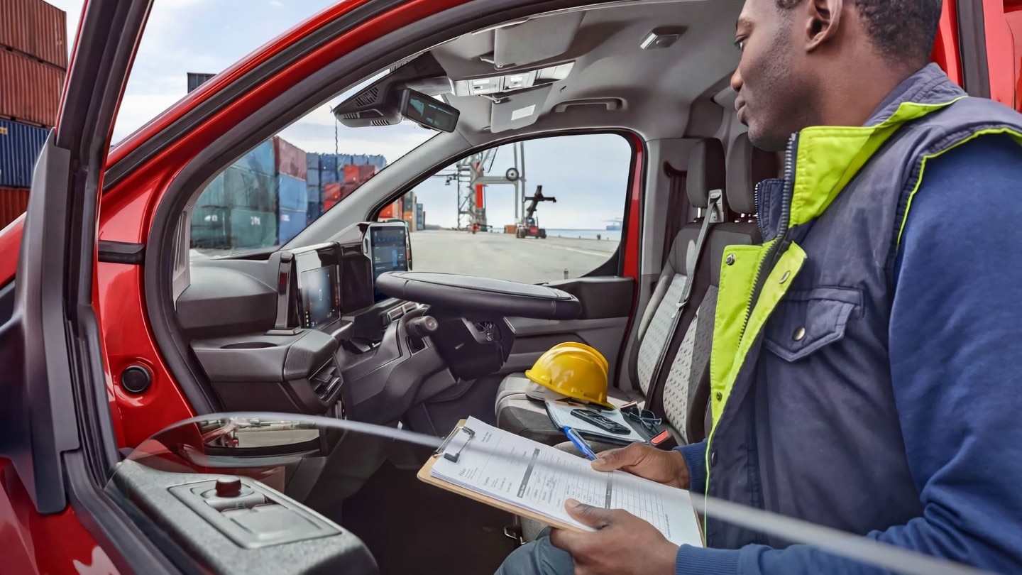 Dans un port de marchandises, un homme installé dans un Ford Transit rouge tient un porte-bloc tout en regardant des conteneurs maritimes et une grue.