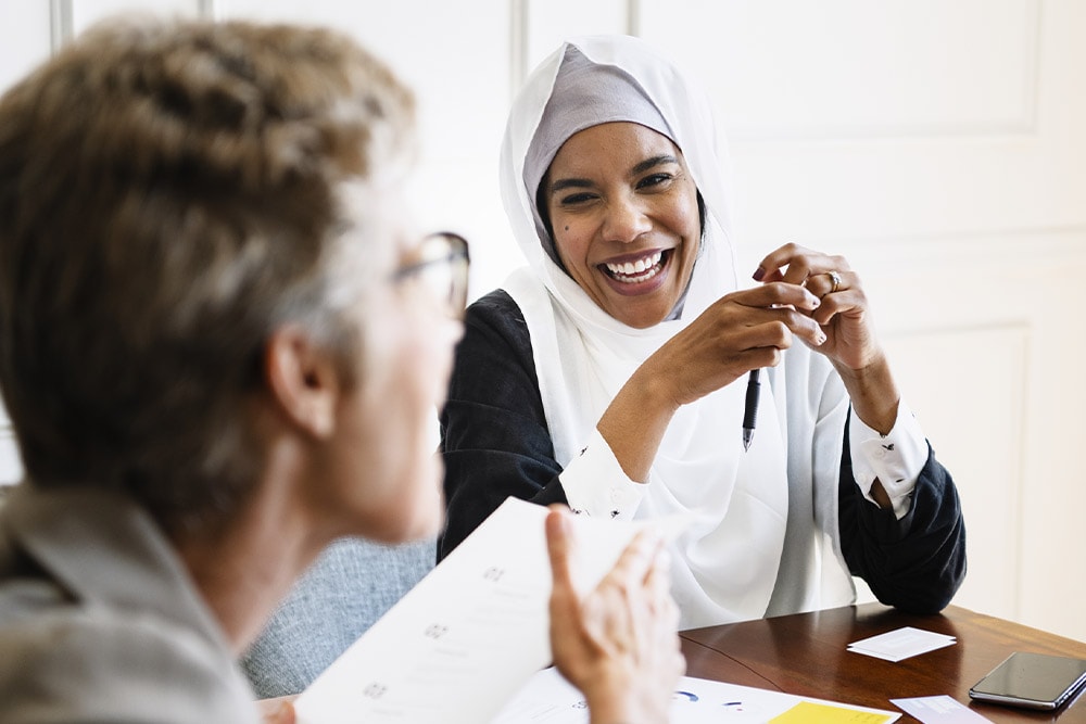 Une femme en hijab blanc sourit lors d'une réunion, tenant un stylo. Des papiers et un téléphone sont sur la table.