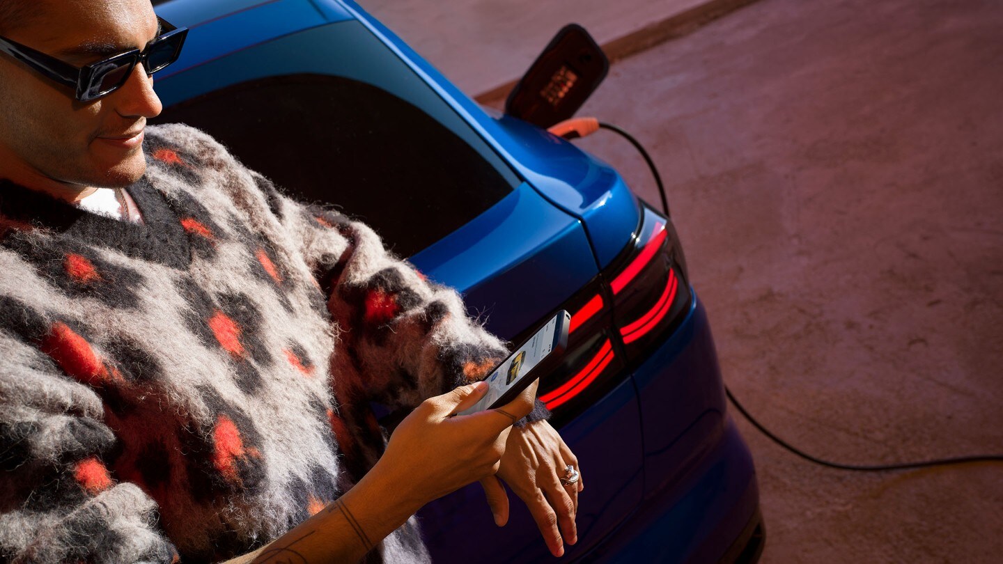 Homme adossé à Ford Capri électrique bleue, regarde téléphone.