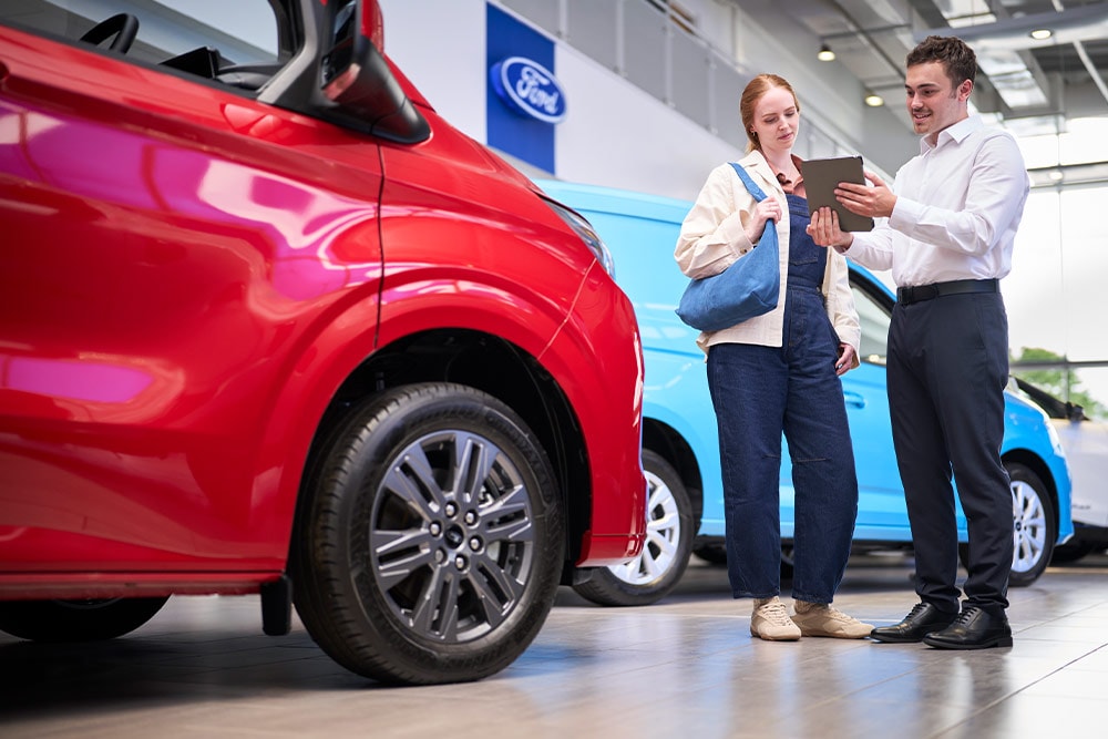 Deux personnes dans un showroom Ford regardent une tablette, avec une fourgonnette Ford rouge au premier plan et une fourgonnette Ford bleue derrière.