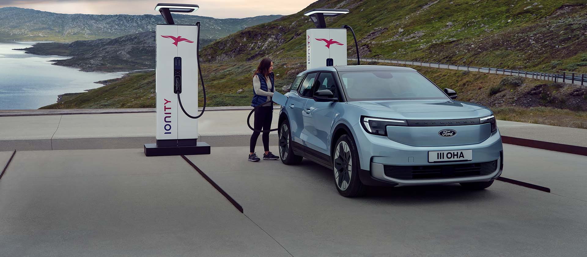 Une femme branche un Ford Explorer bleu 100% électrique, à une borne de recharge publique à l'aide d'un câble.