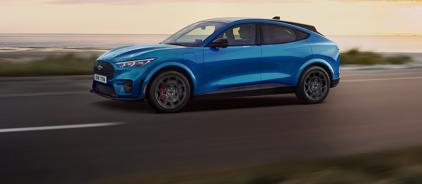 Une Ford Mustang Mach-E électrique bleue roulant le long d'une route côtière au coucher du soleil.