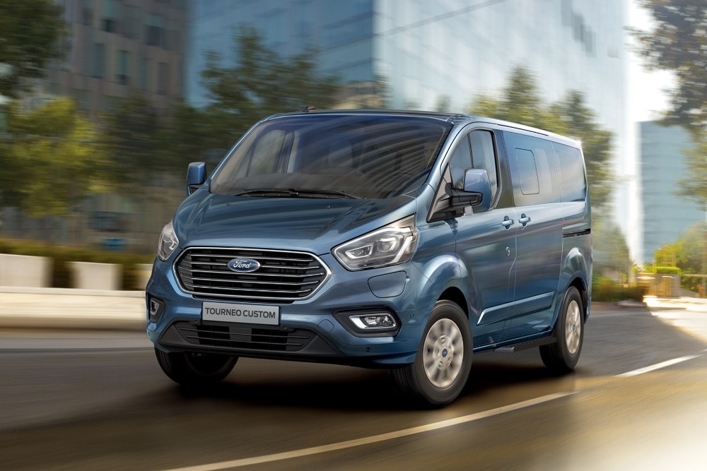 Vue avant trois-quarts d'un Ford Tourneo Custom bleu roulant dans une rue de ville.