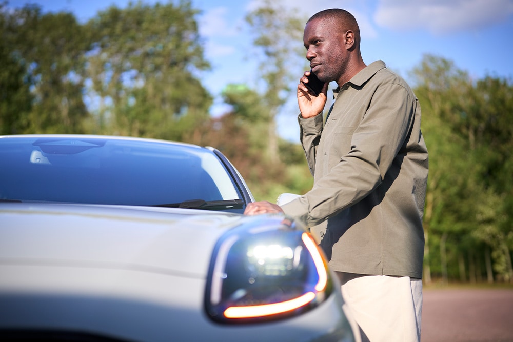 Une personne téléphone à côté d'une voiture de couleur claire en extérieur. 