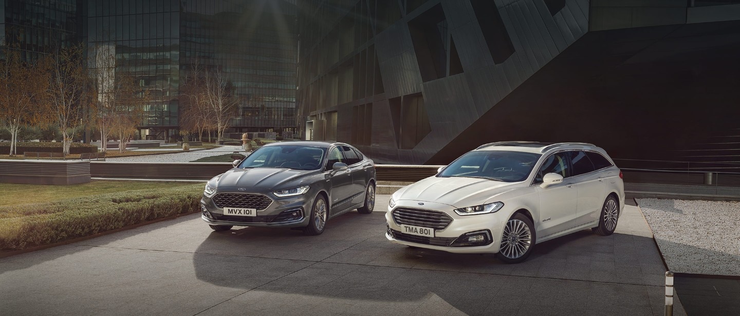 White and dark Ford  Mondeo Titanium HEV and Vignale HEV parking next to modern building frontal view