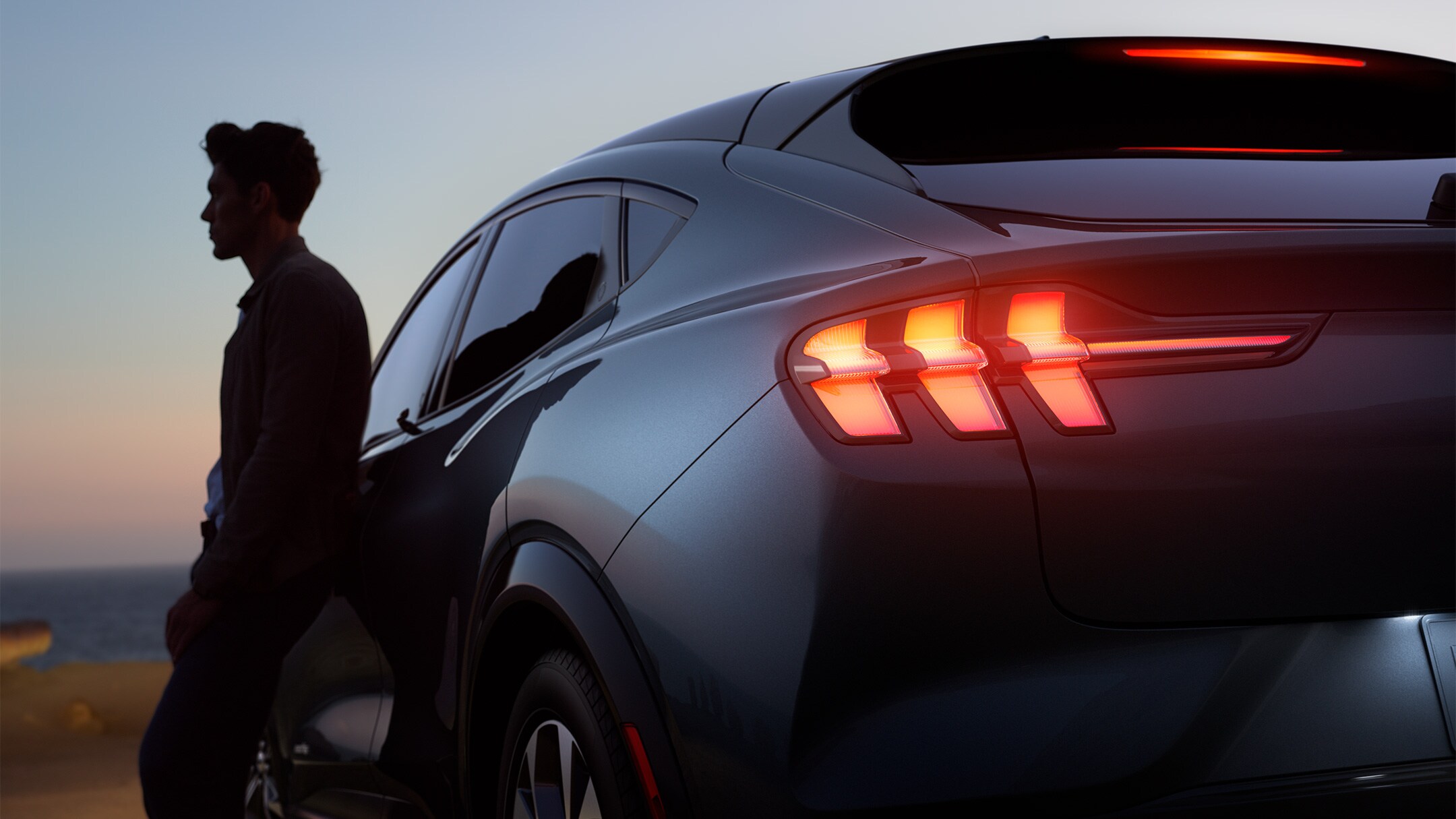 Un homme appuyé sur la porte de sa Nouvelle Ford Mustang Mach-E au coucher du soleil.