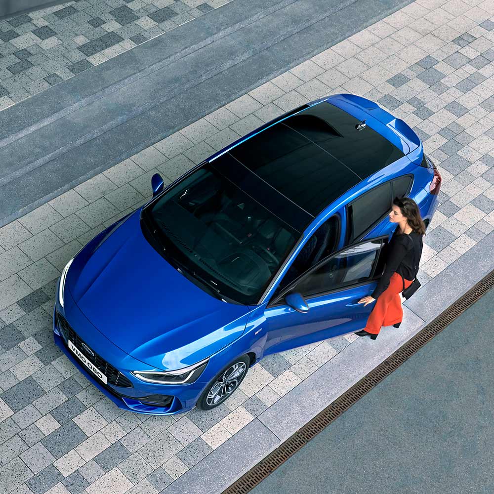 Vue aérienne d'une femme montant dans sa Ford Focus bleue dotée d'un toit panoramique en verre.