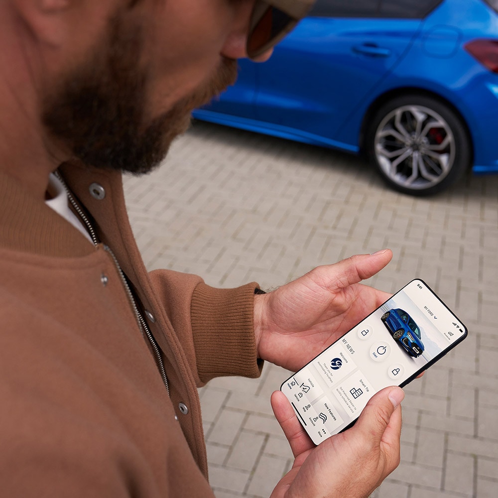 Une personne tenant un smartphone affichant l'application FordPass™, avec une Ford Focus bleue en arrière-plan.