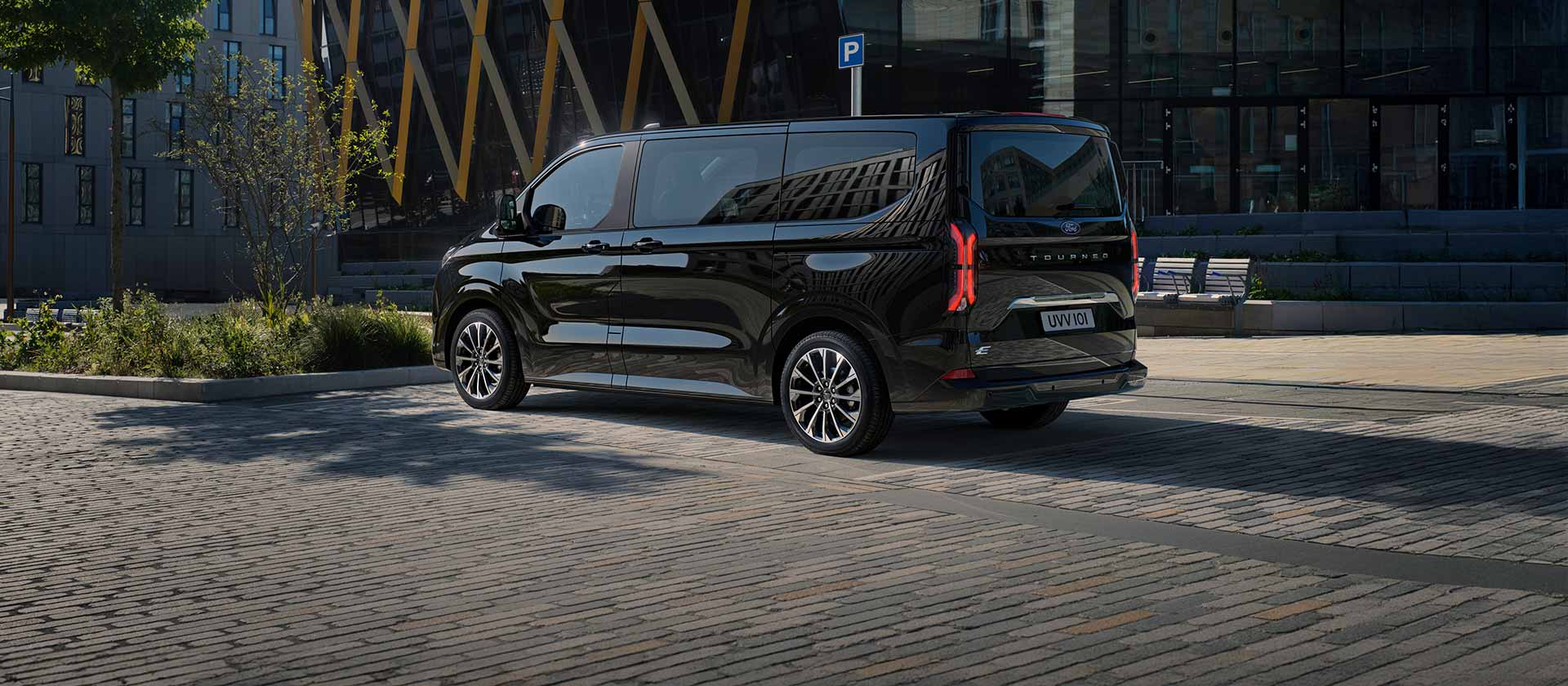 Un Ford E-Tourneo Custom noir est stationné, montrant le côté et l’arrière devant un bâtiment moderne.