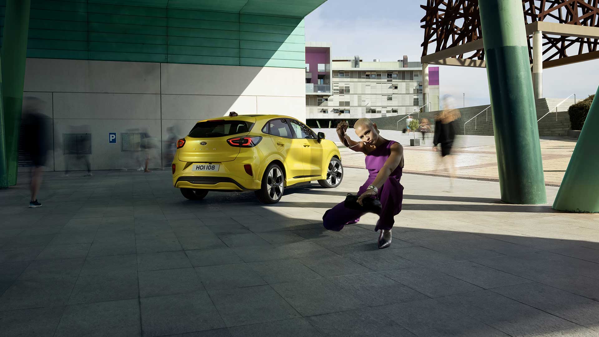 Vue arrière d’une Ford Puma Gen-E® électrique jaune stationnée sous un pont, près d’une personne habillée en violet.