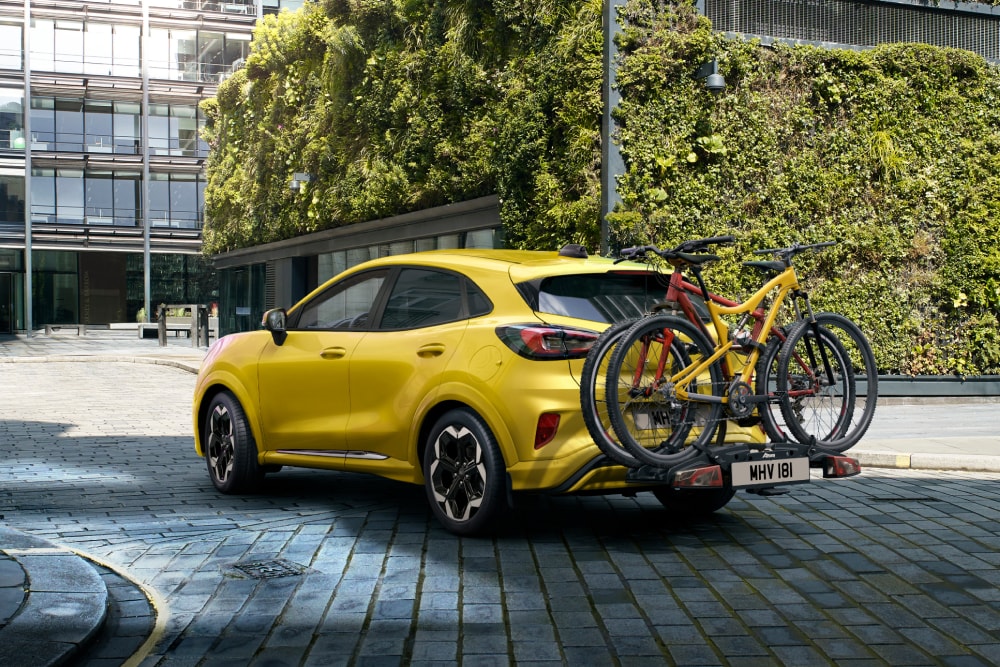 Une Ford Puma Gen‑E jaune garée en milieu urbain, avec deux vélos fixés sur un porte‑vélos arrière.