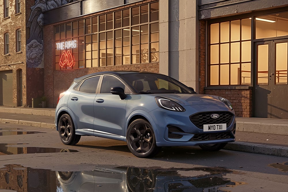 Un Ford Puma bleu garé dans une rue au coucher du soleil, avec des flaques reflétant les bâtiments.