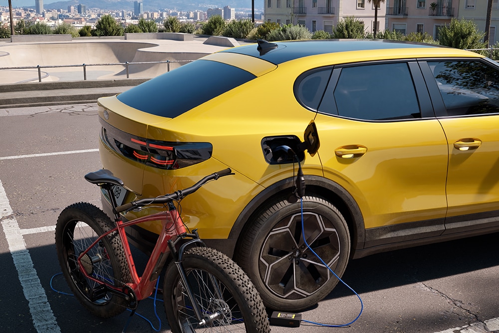 Ford Capri® jaune garée en milieu urbain, rechargeant un vélo électrique rouge via son port de charge.