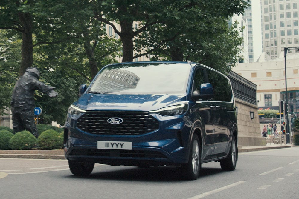Un Ford Tourneo Custom bleu roule dans la rue d'une ville.