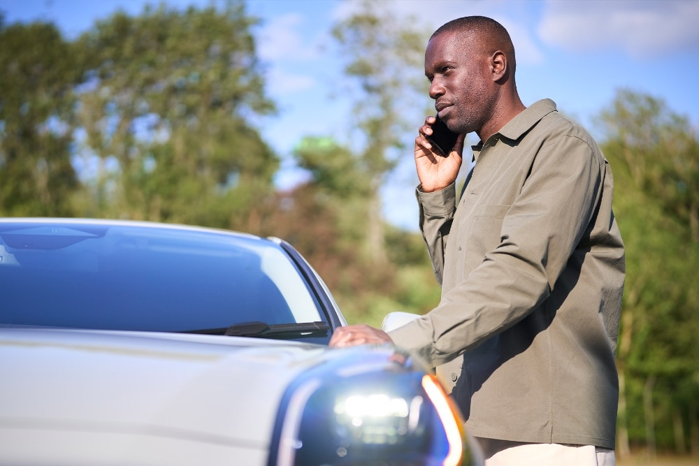 Une personne téléphone à côté d'une voiture de couleur claire en extérieur. 