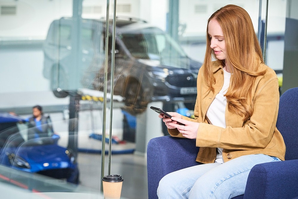 Une personne au téléphone dans l'espace d'attente du service Ford, avec un fourgon sur un pont élévateur et une voiture bleue visibles à travers la baie vitrée.