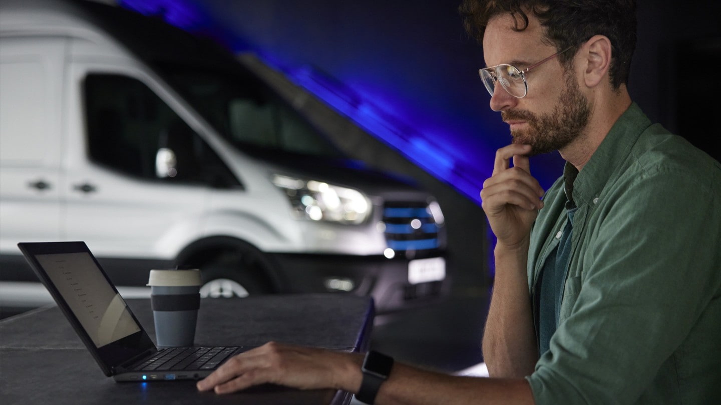 Un homme travaillant sur son ordinateur avec un Ford E-Transit dans le fond.  