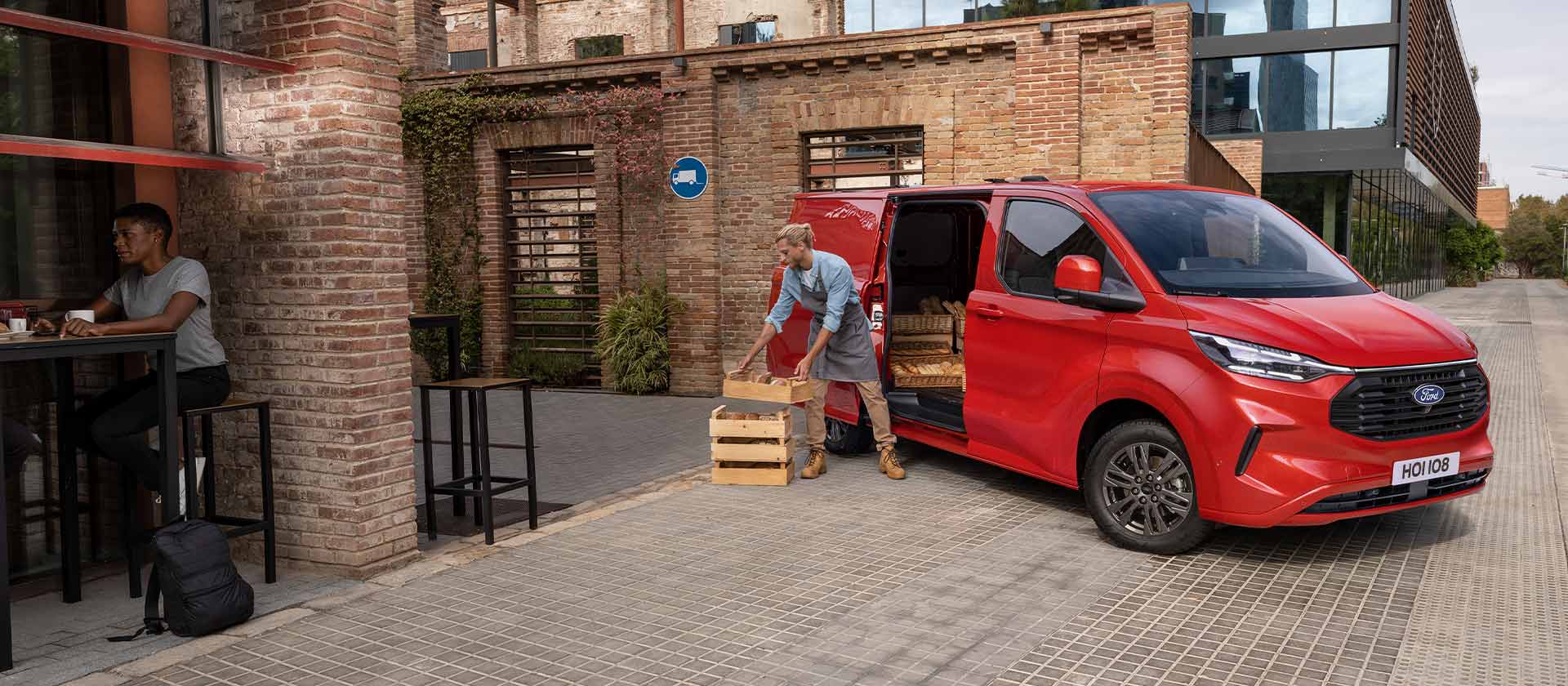 Une personne décharge des caisses d'un fourgon Ford Transit Custom rouge stationné devant un coffee shop. 