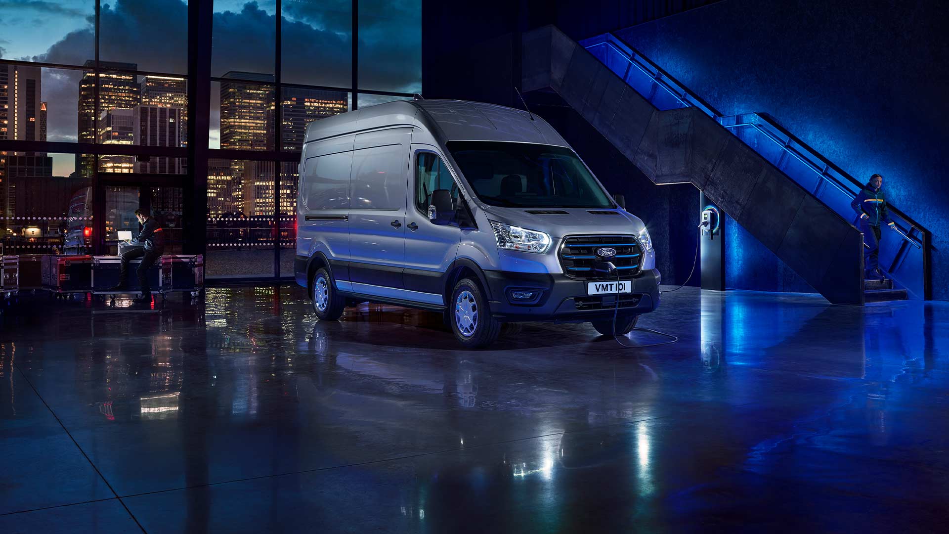 Un fourgon électrique Ford E-Transit argenté est stationné à l’intérieur, avec un horizon urbain visible à travers de grandes fenêtres. 