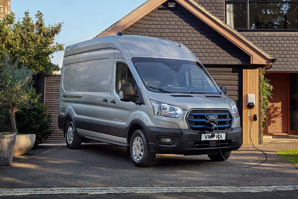 Un fourgon E-Transit Ford argenté est stationné dans une allée bordant une maison et branché à une station de recharge.