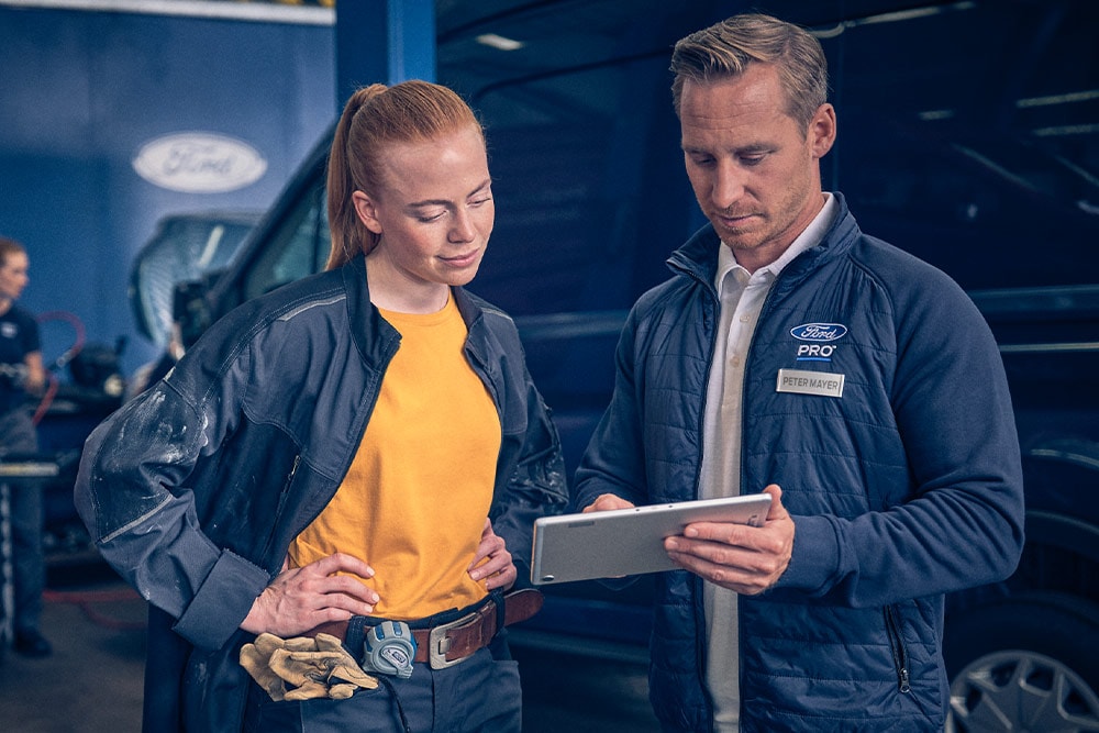 Deux mécaniciens dans un garage regardant une tablette face à un fourgon électrique Ford E-Transit.