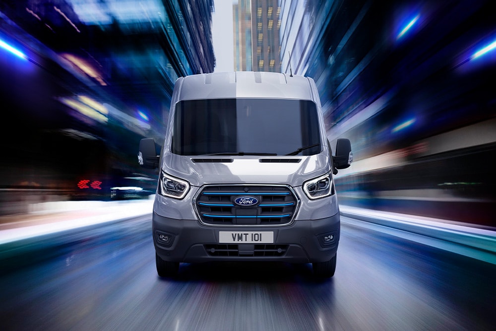 Front view of a grey Ford Transit van with blue grille accents, driving at speed through a city at night.