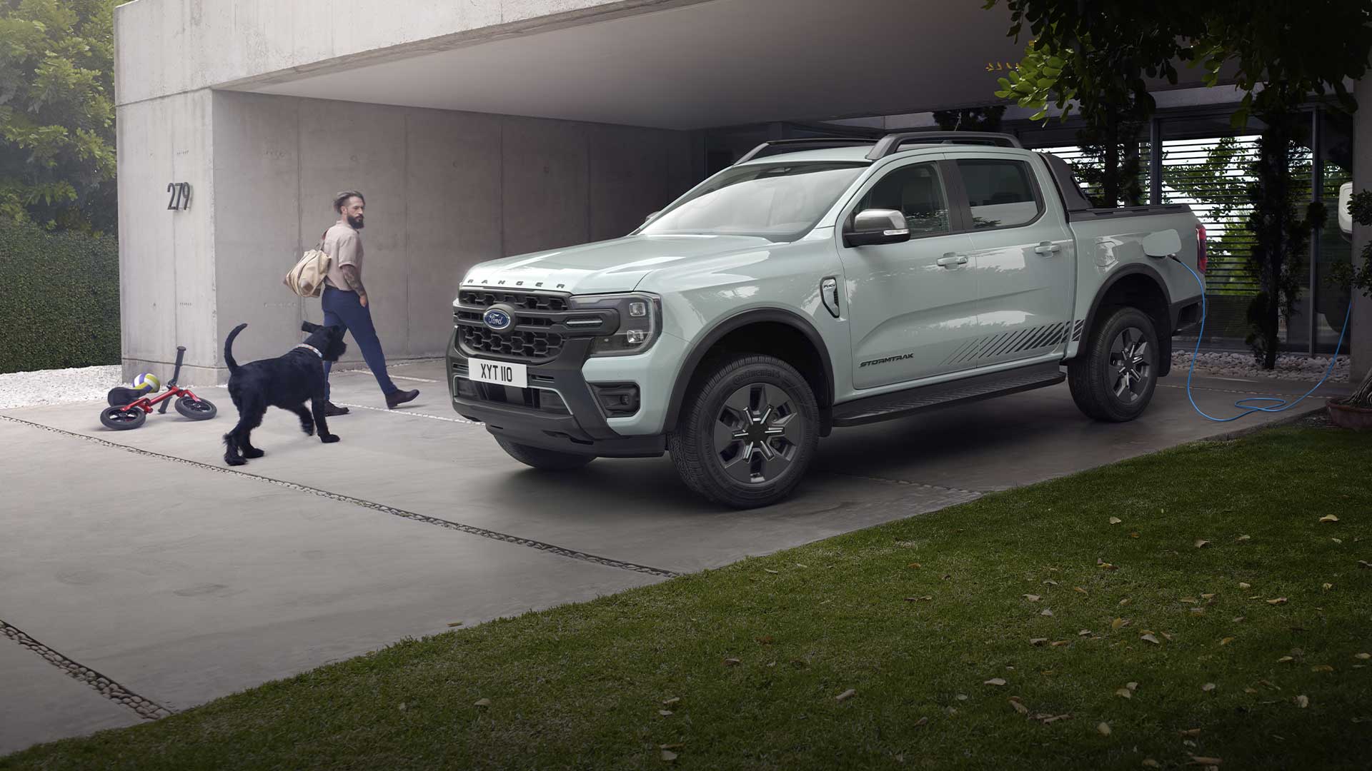 Un homme et son chien s’approchent d’un pick-up Ford Ranger hybride rechargeable gris en train de charger dans une allée. 