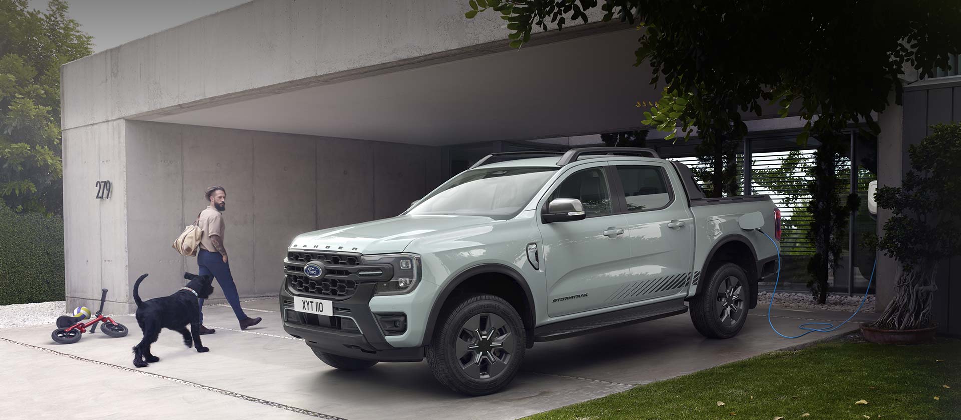 Un homme et son chien entrent dans une maison moderne en passant devant un Ford Ranger Stormtrak PHEV en cours de recharge. 