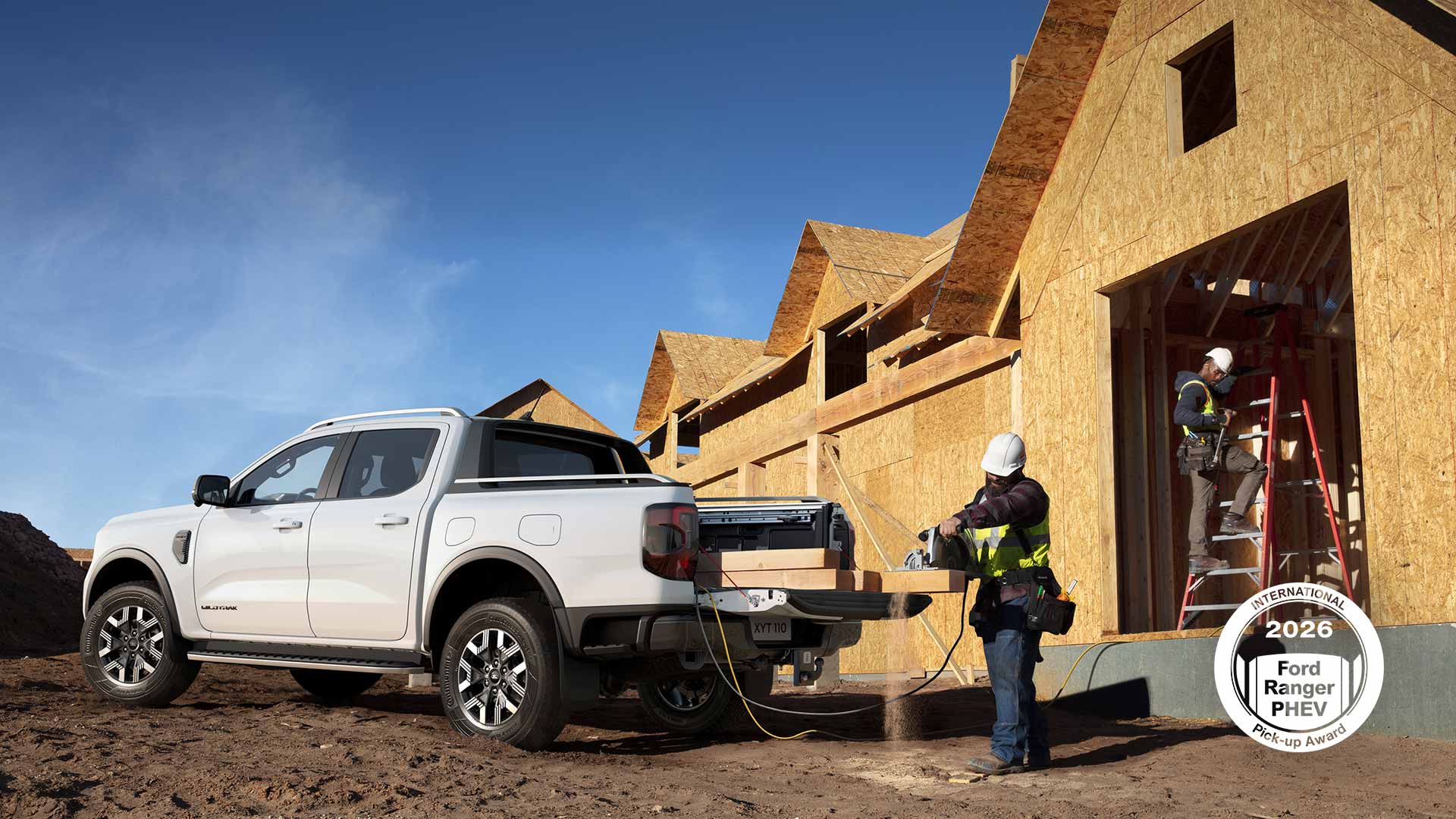 Un pick-up Ford Ranger blanc est stationné sur un chantier. Un ouvrier coupe une planche de bois grâce à une scie alimentée par le véhicule.