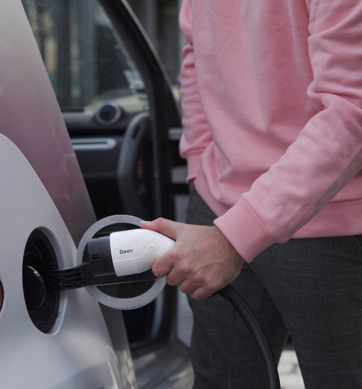 Une personne en pull rose branche un câble de recharge dans la prise d'un véhicule électrique.