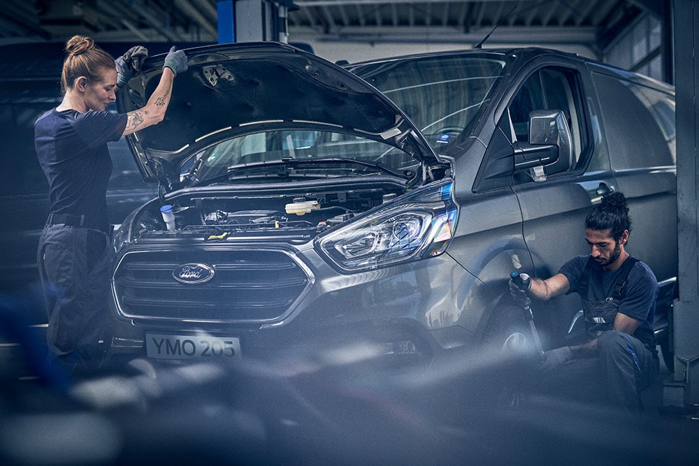 Deux techniciens travaillent sur un Ford Transit Custom gris dans un garage. L'un inspecte le moteur, l'autre la roue avant.