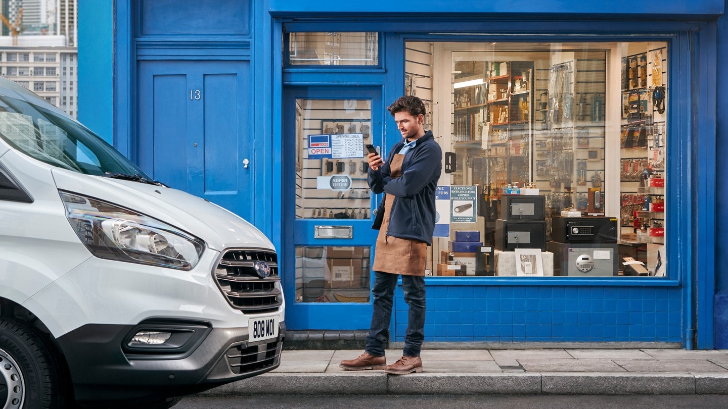 Une personne devant un magasin consultant son téléphone portable et un fourgon Ford garé à côté du magasin. 