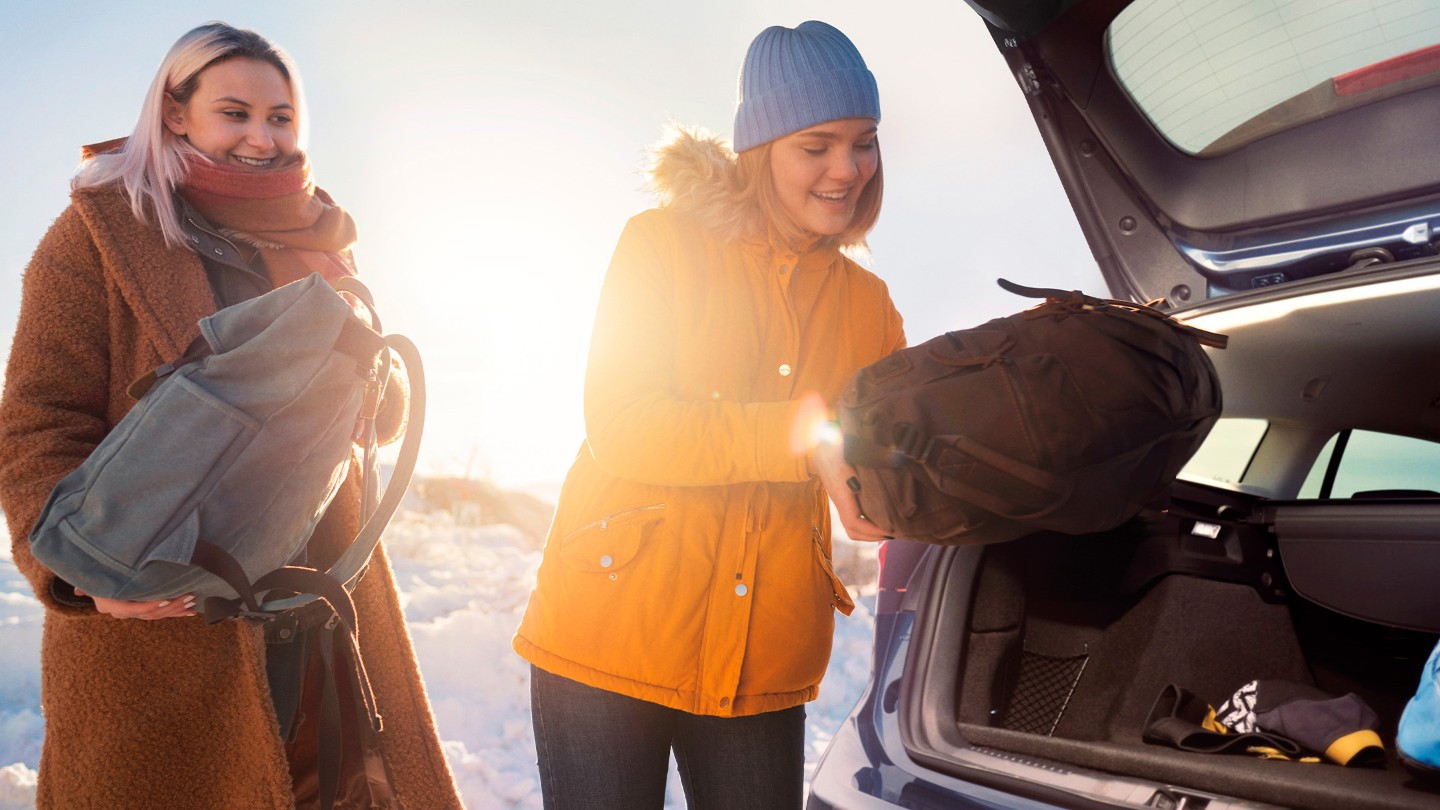 Une femme souriante en vêtements d’hiver qui charge des sacs dans le coffre d’une voiture dans un paysage enneigé