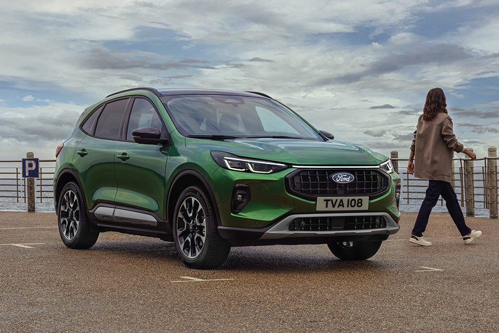 Front 3/4 view of a green Ford Kuga parked by a coastal railing, with two people nearby.
