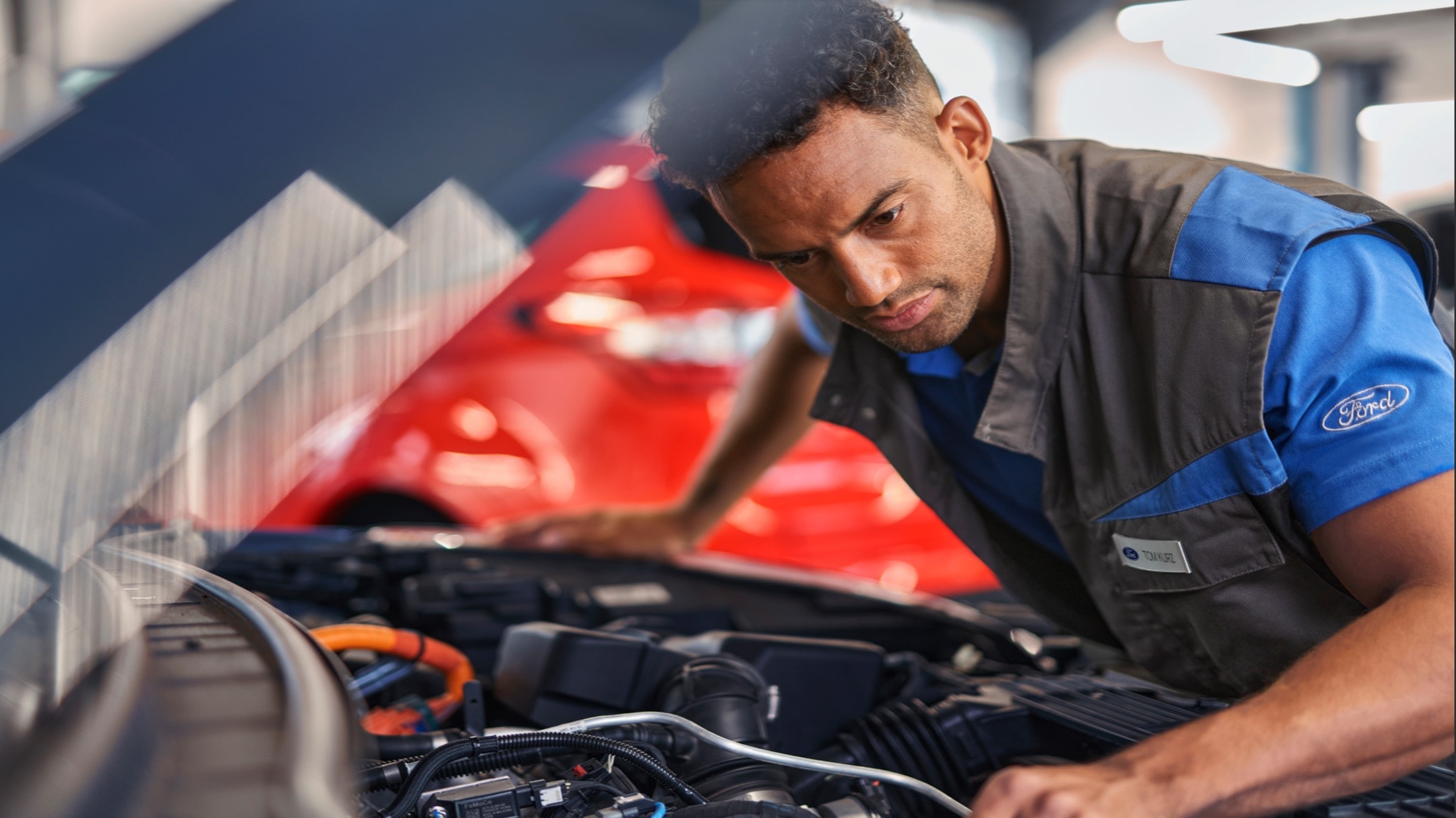 Mécanicien Ford inspectant le moteur d’un véhicule dans un atelier, portant un uniforme bleu et noir avec le logo Ford